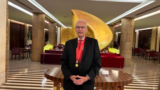 Älterer Mann mit Brille und rotem Anzug, trägt eine Medaille um den Hals, steht in einem eleganten Restaurant vor einer goldenen Skulptur.
