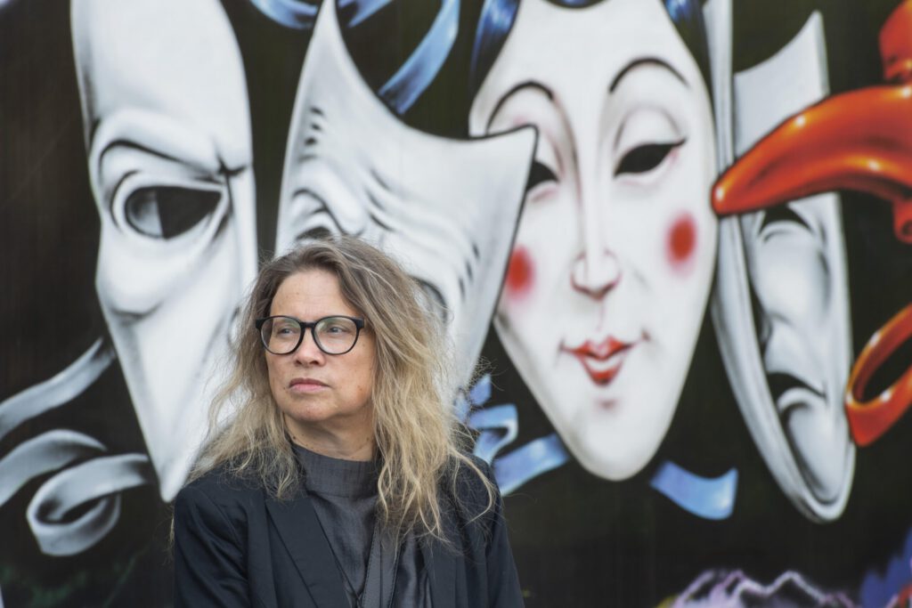 Frau mit langen, lockigen Haaren und Brille steht vor einer Wand mit bunten Masken und Gesichtern.
