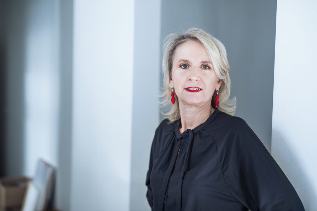 Frau mit blonden Haaren und roten Ohrringen, trägt ein schwarzes Oberteil, steht in einem modernen Raum.