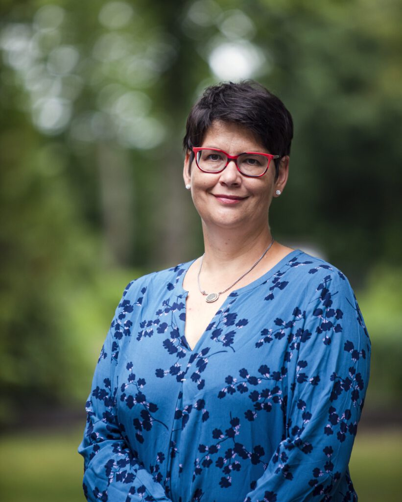 Frau mit kurzen, dunklen Haaren und Brille, trägt ein blaues Kleid mit floralem Muster, lächelt freundlich im Freien.