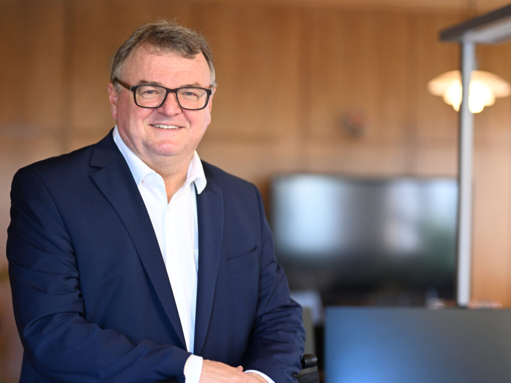 Mann in Anzug mit Brille, lächelt freundlich, steht in einem modernen Büro mit Holzinterieur.