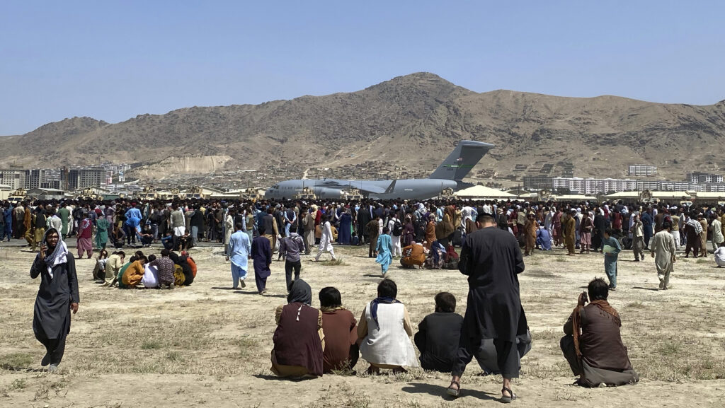 Große Menschenmenge auf einem Feld in Kabul, Afghanistan, vor einem Militärflugzeug, umgeben von Bergen und städtischer Architektur.