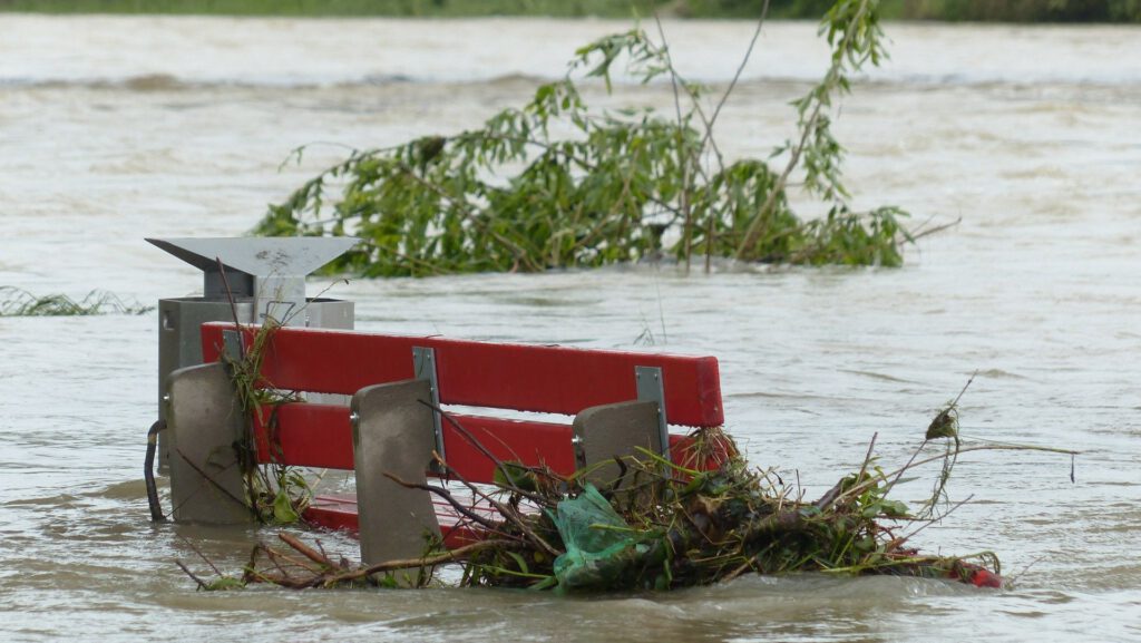 Eine rote Bank steht teilweise im Wasser, umgeben von Treibgut und Ästen, das durch eine Überschwemmung verursacht wurde.