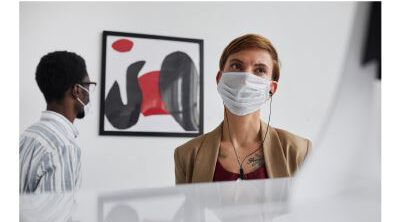 Zwei Personen in einem modernen Raum, eine Frau mit kurzem, rotem Haar und Mundschutz, die auf ein Kunstwerk schaut, während ein Mann im Hintergrund mit Brille und ebenfalls Mundschutz steht.