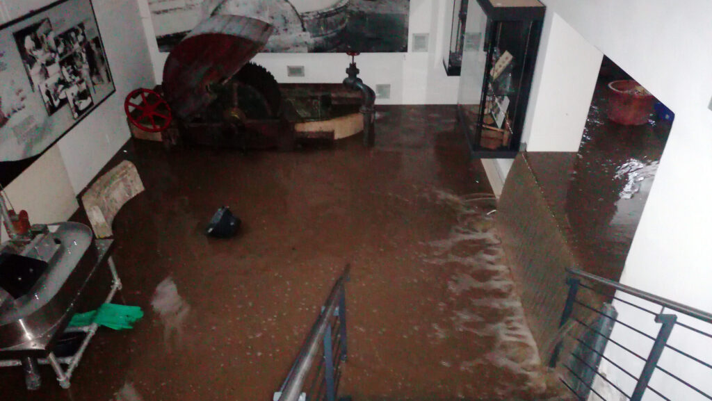 Überfluteter Raum mit Wasser auf dem Boden, Möbel sind teilweise beschädigt. Sichtbare Wasserstände an Wänden und Treppe.