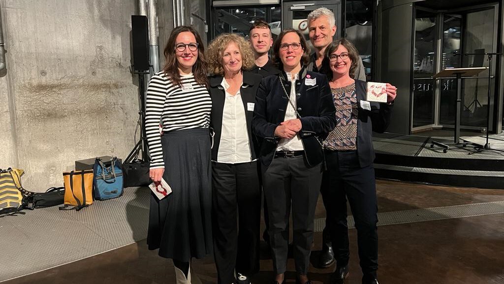 Gruppe von sechs Personen in einem modernen Veranstaltungsraum, lächelnd und posierend für ein Foto. Einige tragen Namensschilder. Von links nach rechts: Gülsah Stapel, Dr. Claudia Emmert, Dominik Busch, Dr. Felicia Sternfeld, Prof. Dr. Joachim Baur, Dr. Alina Gromova
