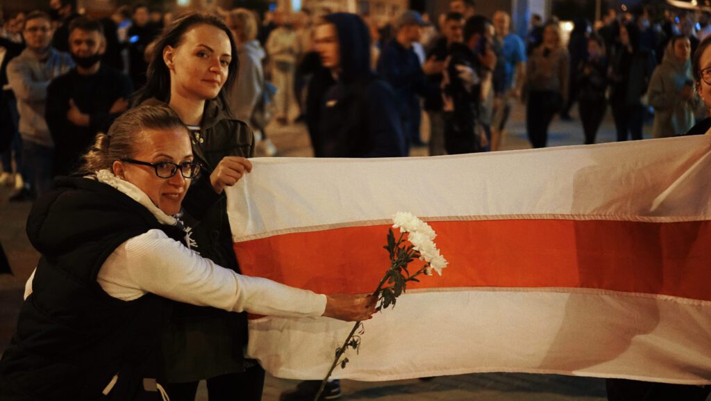 Zwei Frauen halten eine große weiß-rote Flagge und eine Blume in einer Menschenmenge während einer Demonstration.