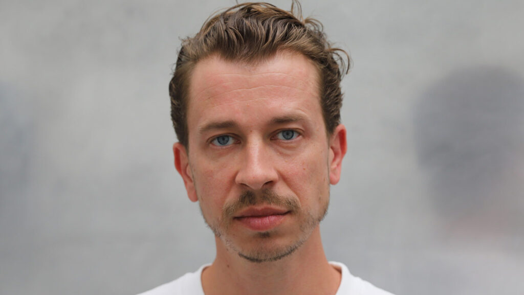 Mann (Dominik Busch) mit kurzen, lockigen Haaren und blauen Augen, trägt ein weißes T-Shirt, schaut direkt in die Kamera.