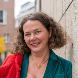 Eine weibliche Person lehnt an einer Mauer, trägt grünes Sakko, weiße Bluse, schwarze Hose und hält rotes Jackett über die Schulter.