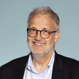 Ein Mann mit grauem Haar und Bart trägt ein hellblaues Hemd und ein schwarzes Sakko vor einem einfarbigen hellblauen Hintergrund.