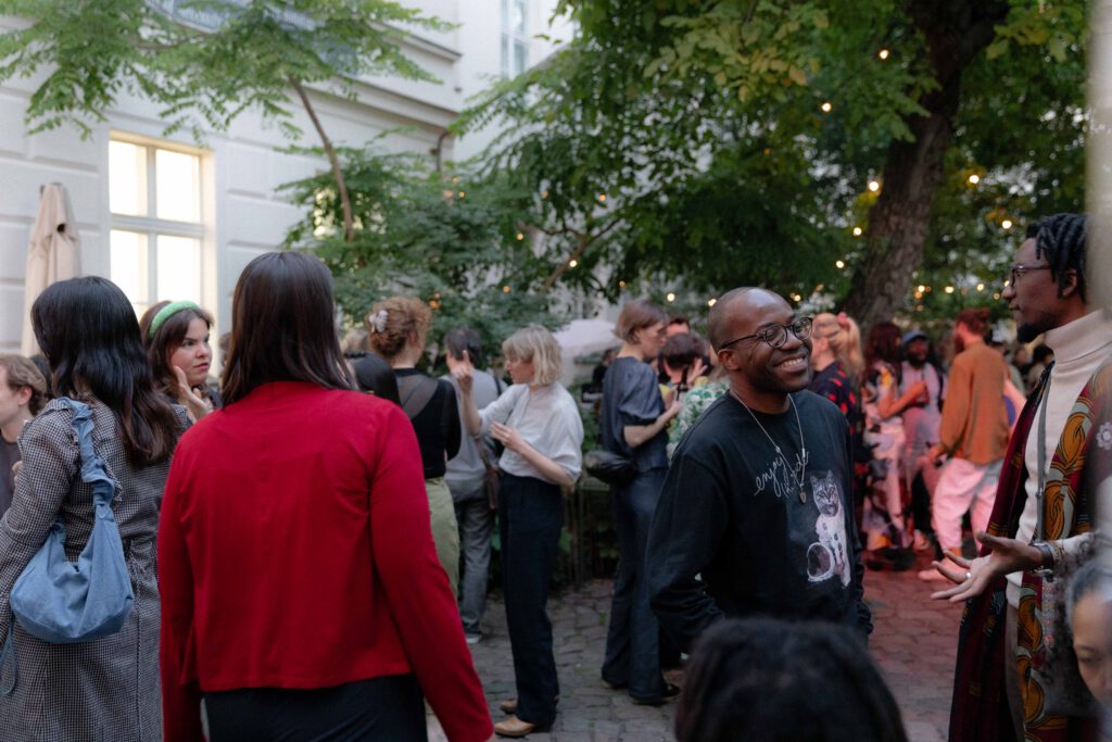 Menschen in einem Innenhof bei Dämmerung, einige unter Bäumen mit Lichterketten, die meisten stehen und unterhalten sich