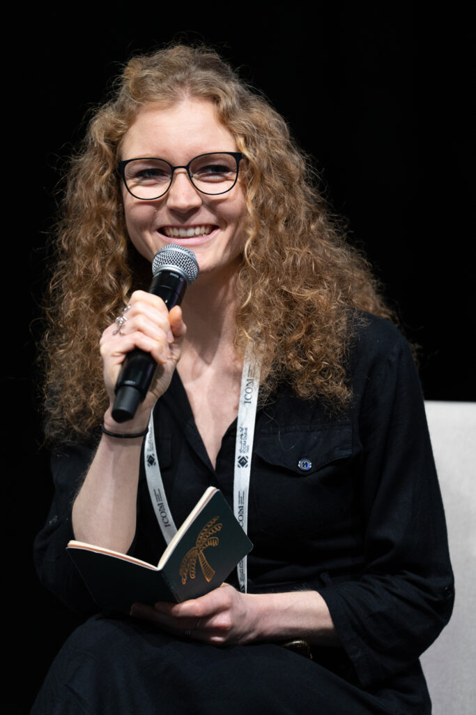 Frau mit lockigen Haaren, einem Mikrofon und einem Notizbuch in der Hand.