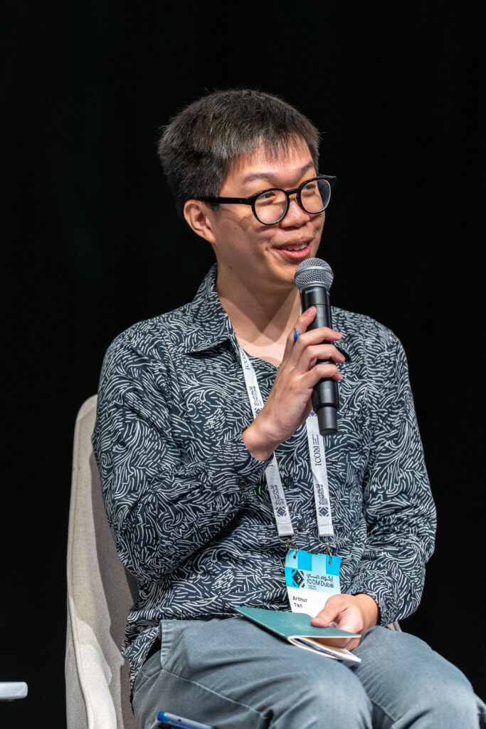 Person mit Brille und gemustertem Hemd sitzt auf Stuhl und hält Mikrofon und Notizbuch in der Hand