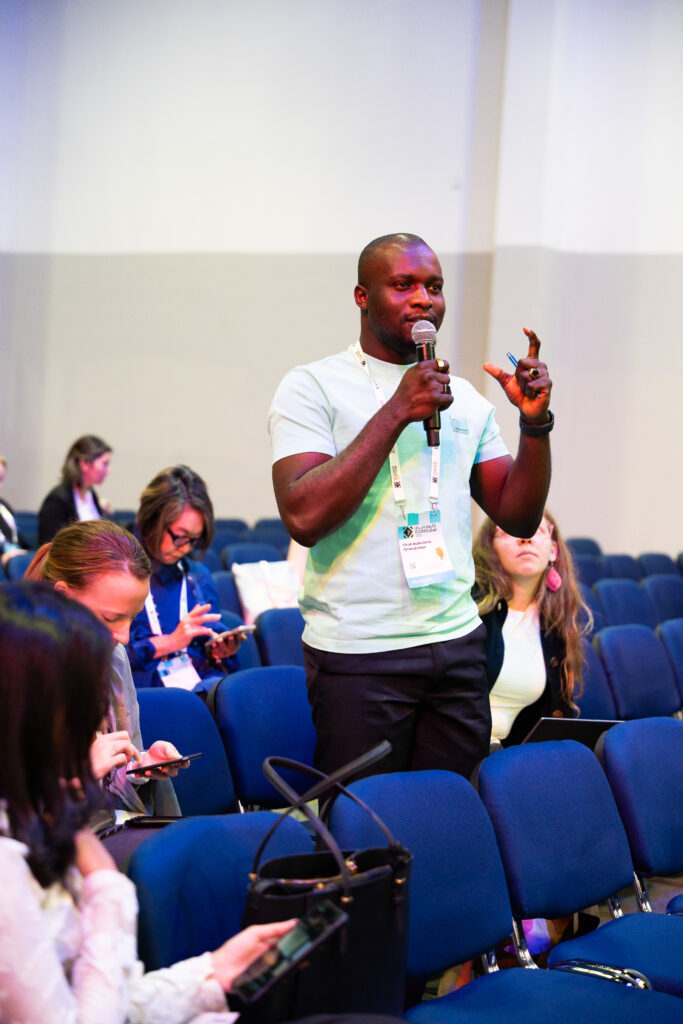 Mann steht in einem Saal mit blauen Stühlen und hält ein Mikrofon in der Hand