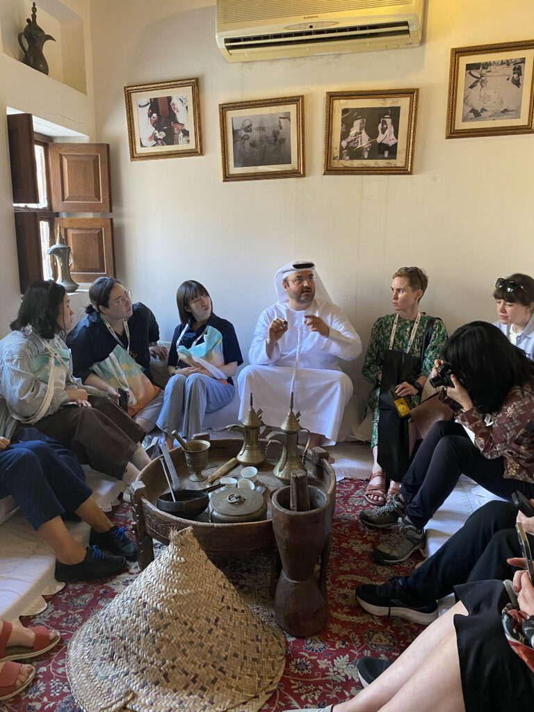 Gruppe von Menschen sitzt im Kreis auf Sitzkissen in einem Raum mit traditionellen Teekannen und einem großen geflochtenen Korb auf einem Teppich