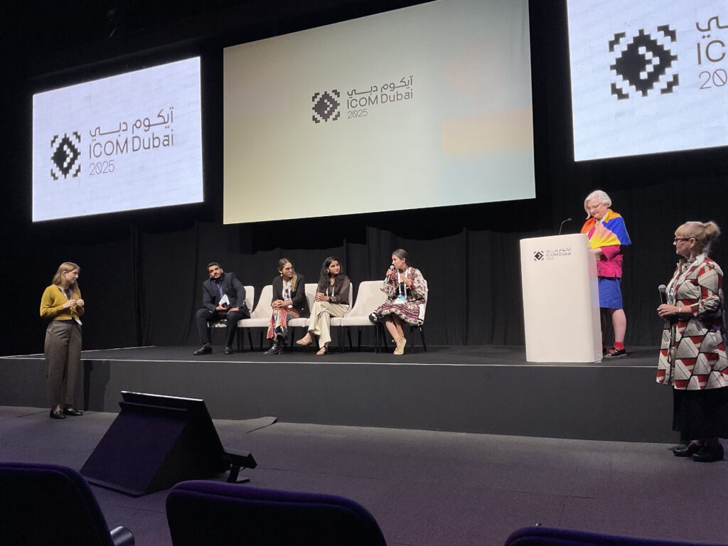 Podiumsdiskussion mit fünf Personen auf einer Bühne vor zwei Bildschirmen mit dem Logo und Schriftzug 'ICOM Dubai 2025', eine Frau am Rednerpult und zwei weitere Frauen stehen seitlich auf der Bühne