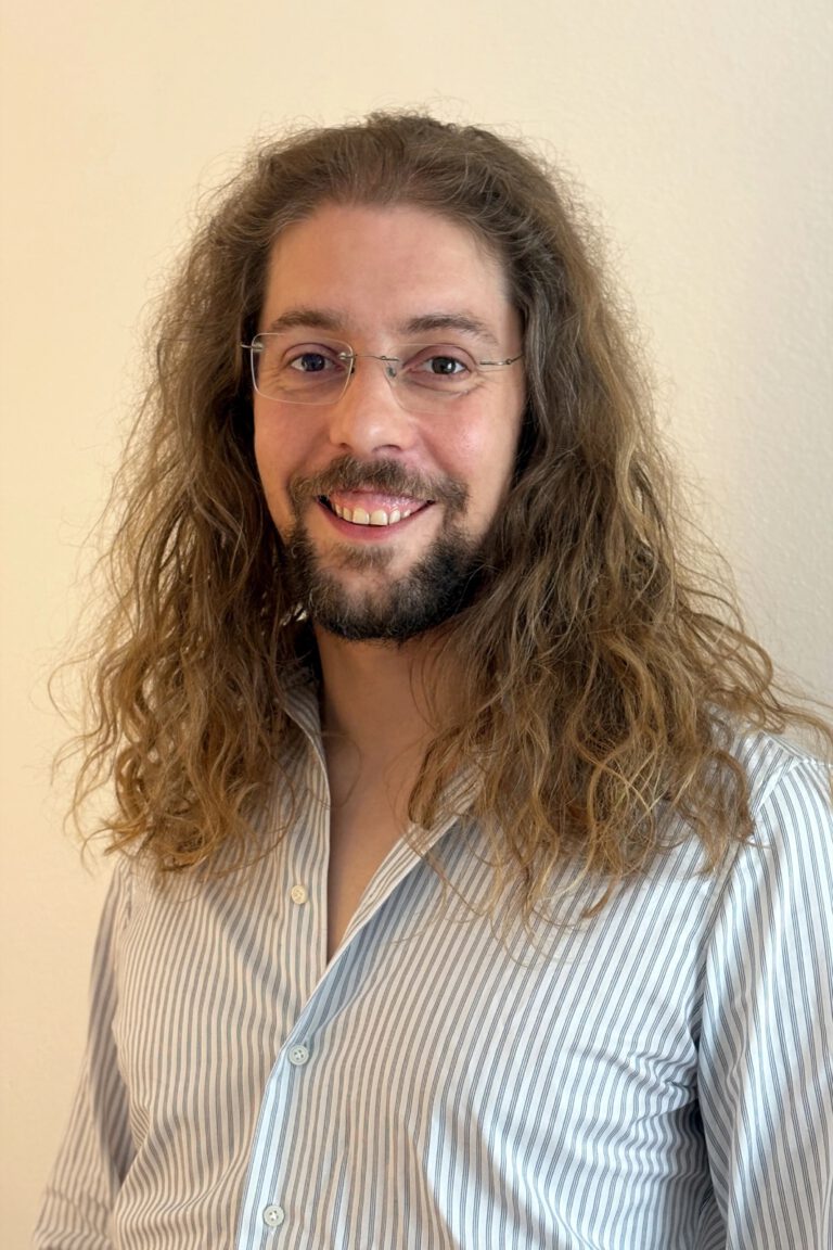 Lächelnde Person mit langem, lockigem Haar, Bart und randloser Brille trägt ein gestreiftes Hemd vor hellem Hintergrund.