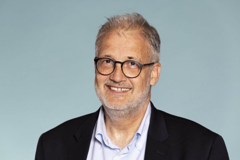 Lächelnde Person mit kurzem, grauem Haar, Bart und runder Brille trägt ein dunkles Sakko und ein hellblaues Hemd vor blaugrauem Hintergrund.