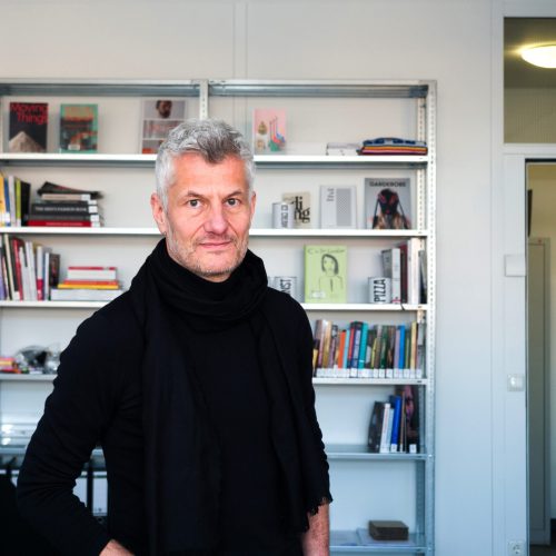 Prof. Dr. Joachim Baur, Foto: Silke Wawro Mann (Prof. Dr. Joachim Baur) mit grauen Haaren und Bart, trägt schwarzen Pullover, sitzt vor einem Bücherregal mit verschiedenen Büchern.