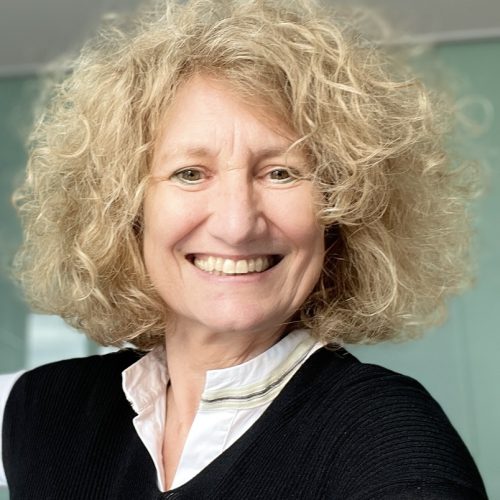 Frau mit lockigem, schulterlangem Haar trägt eine schwarze Strickjacke über einer weißen Bluse mit Stehkragen.