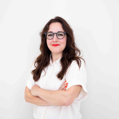 Frau mit langen, gewellten Haaren trägt ein weißes Hemd und steht mit verschränkten Armen vor weißem Hintergrund.